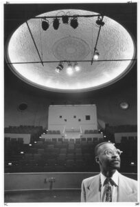 a black and white photo with 1993 director Steve Sneed in the foreground looking off camera to the right with theater seating expanding out behind him.