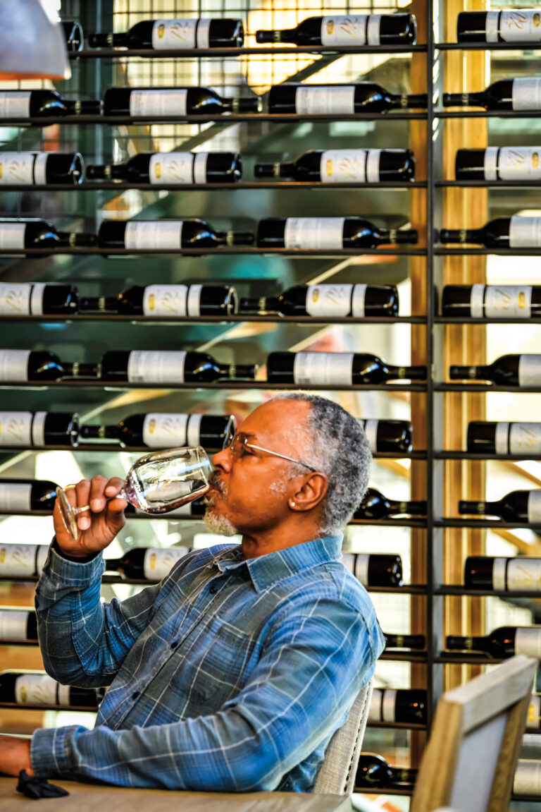 The profile of an older black man sipping a glass of wine with an artful display of bottled wine in the background.