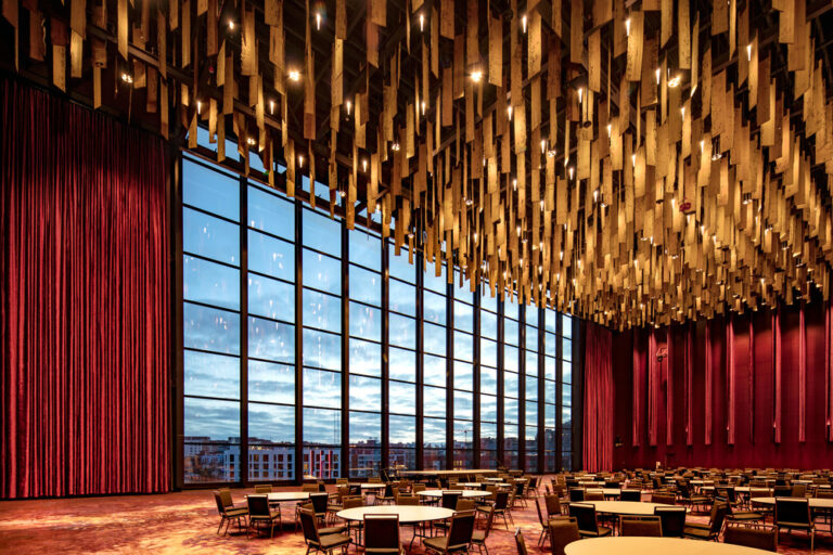 Convention center ballroom with dark red fabric draping, suspended wooden planks, chairs, tables, and floor to ceiling windows