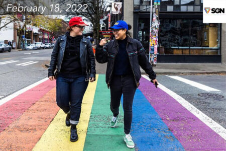 Two people walking across a rainbow colored crosswalk in the city.