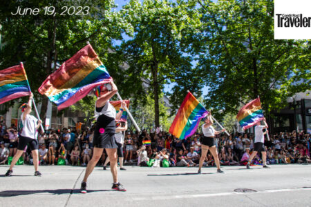Pride parade with a crowd watching performers walking with the Pride flag.