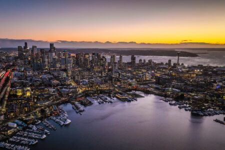 Aerial image of the Seattle skyline at dusk, looking southwest from above Lake Union. The dominant color is purple, accented by golden orange on the horizon.