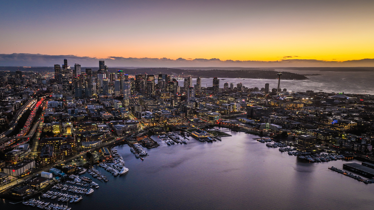 Aerial image of the Seattle skyline at dusk, looking southwest from above Lake Union. The dominant color is purple, accented by golden orange on the horizon.