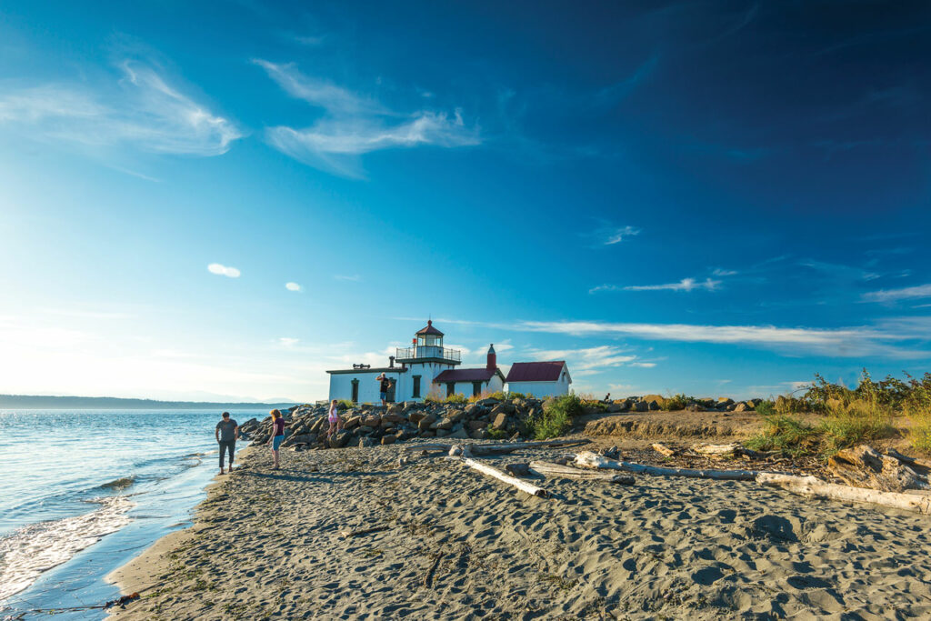 A white lighthouse with a red roof is surrounded by brown sand, driftwood, and green grass. People wearing summer clothes stand on the beach and on the rocks near the lighthouse. The blue Puget Sound water sparkles on the left.