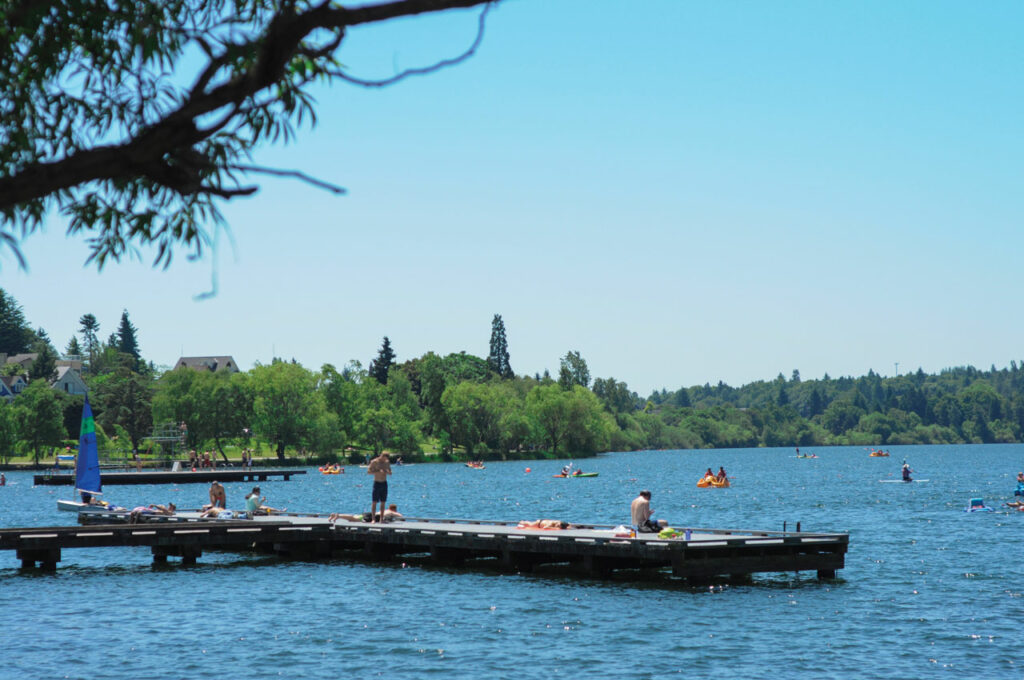 A photo of Green Lake showing a dock sticking out into the water where people are standing and laying down. People in yellow peddle boats and on standup paddle board dot the blue water. Green trees line the background.