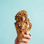A hand holding a waffle cone filled with two scoops of ice cream drizzles with chocolate and peanuts.