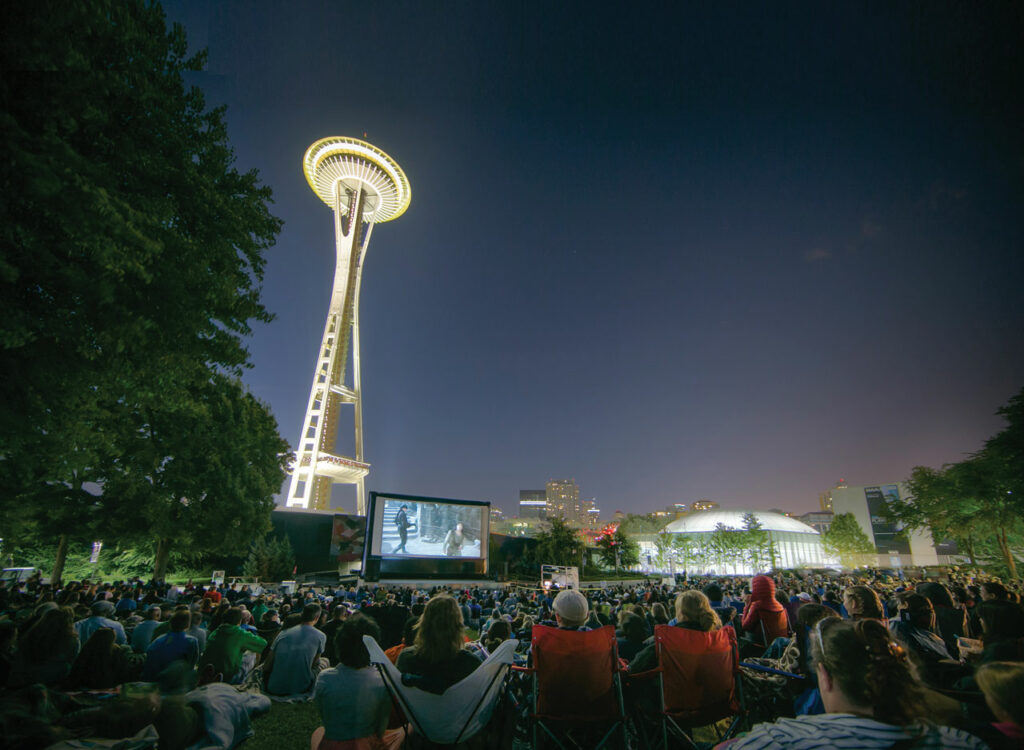 A photo of Movies at the Mural at Seattle Center. The Space Needle is lit up in the background. A crowd of people sit on a lawn in camping chairs and watch a movie on large projector screen.
