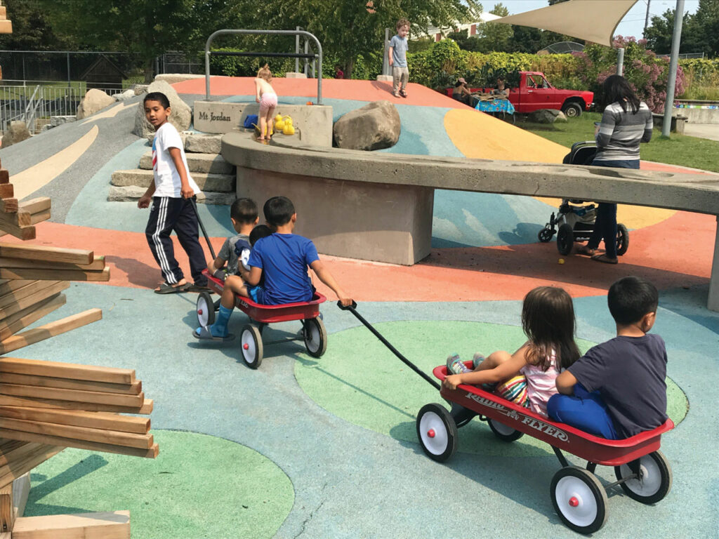 A photo of five kits on a playground that has green, blue, and orange flooring. One kid in a white shirt and black track pants pulls a red wagon with two children sitting it in. A kid in the wagon pulls an additional red wagon with two more kids in it.