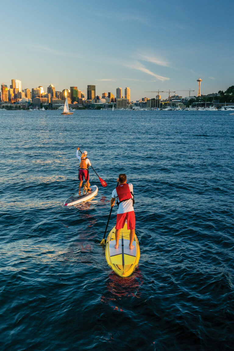 two people wearing while long-sleeve shirts, red shorts, and red life jackets stand on paddle boards in the middle of blue Lake Union. The Seattle skyline and boats are seen in the background.