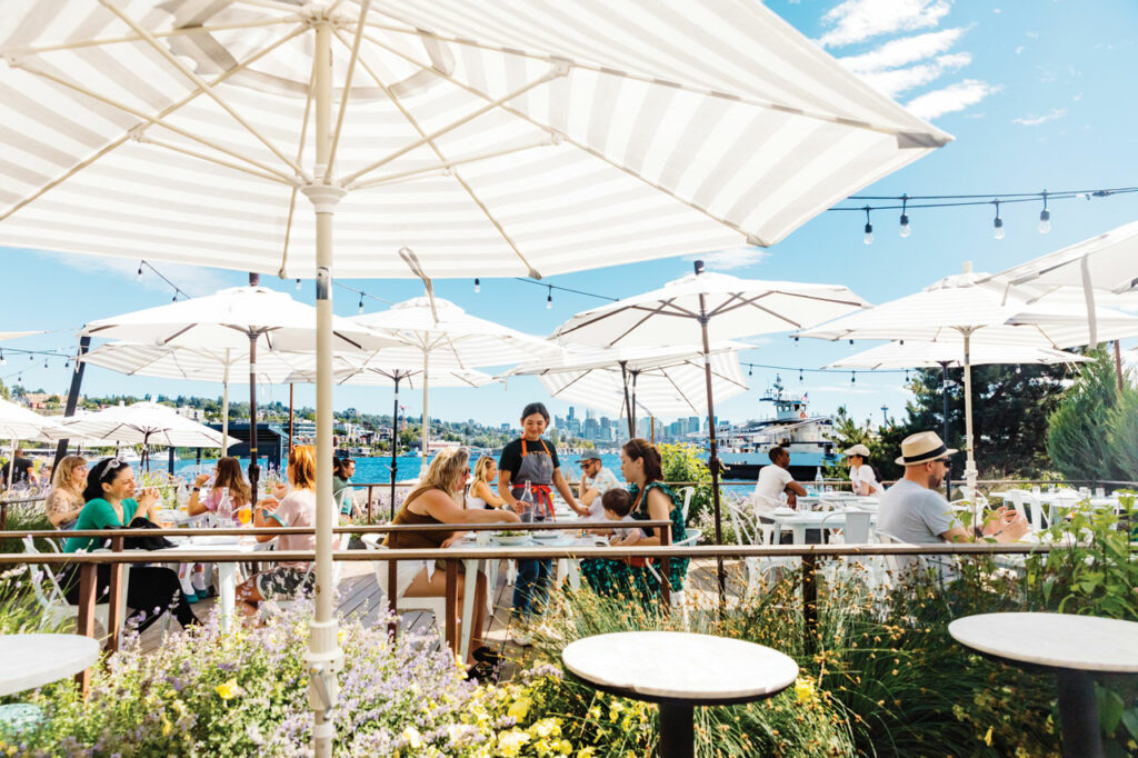 An outdoor patio at a restaurant with white and tan striped umbrellas. People sit at tables on the patio. The water of Lake Union can be seen in the background.