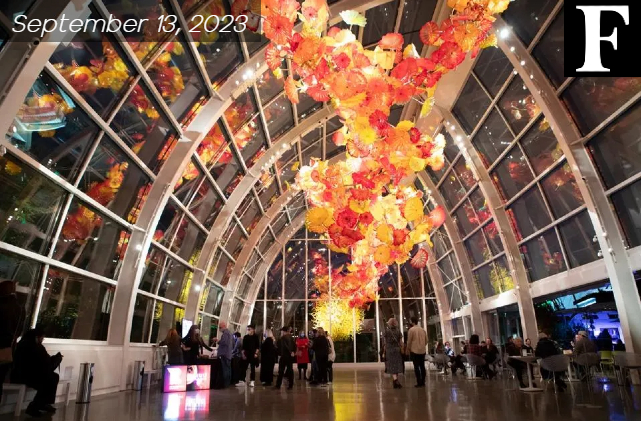 Inside the Chihuly Garden and Glass museum. Hanging orange colored glass flower shaped design from the roof. Glass windows surrounding the room.