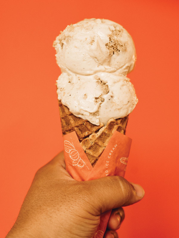 A brown hand holding a waffle cone wrapped in orange paper with two scoops of white ice cream against an orange backdrop
