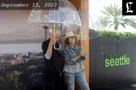 Two middle-aged male and female standing under a clear umbrella with rain coming down.