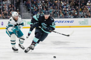 A photo of a Kraken hockey game. Three hockey players are shown in action on the ice. Two are in navy and aqua blue uniforms. One is in a white and teal uniform. The crowd is seen in the stands behind the ice.