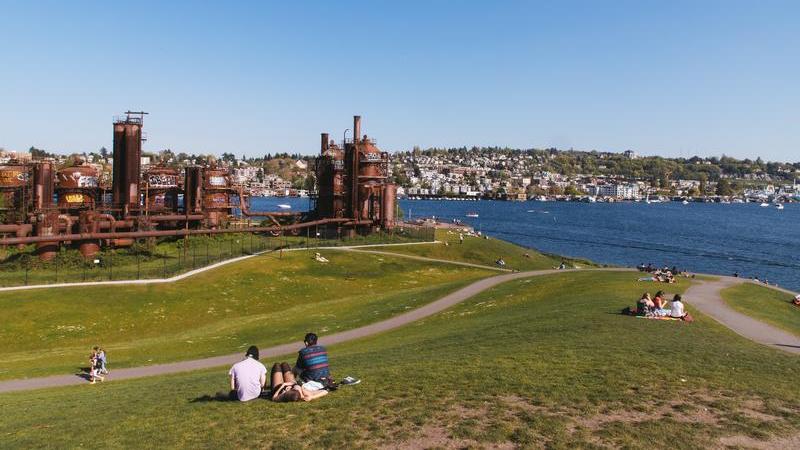 groups of people lounging in the grass with the steampunk-esque towers of Gas Works Park rise in the distance, the waters of Lake Union beyond.
