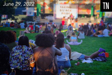 A crowd of people sitting on a lawn in front of a stage with a screen that reads 