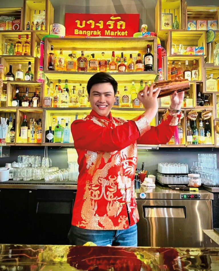 A bartender in a red tunic holding a cocktail shaker. Yellow-lit shelves of alcohol and a bar top are in the background.