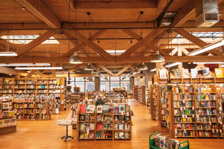 The interior of Elliott Bay Book Company showing several book shelves filled with colorful books. Large wood beams run across the ceiling. A wall of windows is seen in the back of the room.