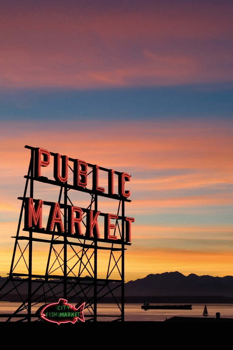 A sunset photo of the Public Market sign at Pike Place Market. The Olympic Mountains and Puget Sound can be seen in the background.