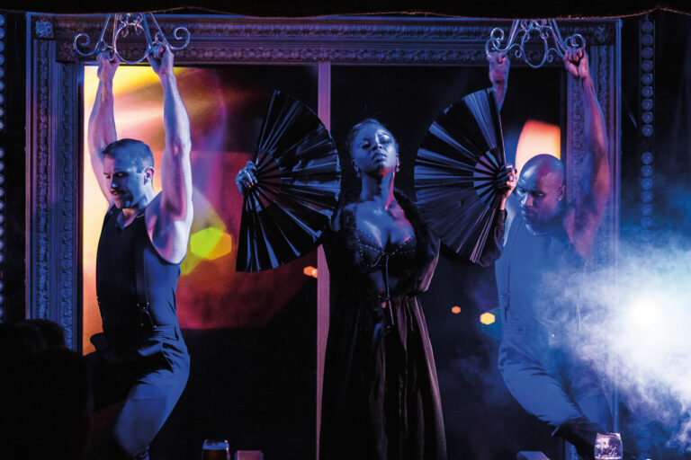 A photo of cabaret dancers wearing black costumes. The person on the left holds two large black fans.