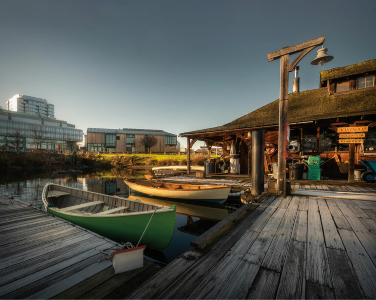A photo of The Center for Wooden Boats showing a dock with one green and one yellow row boat tired up. A wooden building with supplies sitting outside and a sign that says "boat shop" is seen in the back right of the photo.