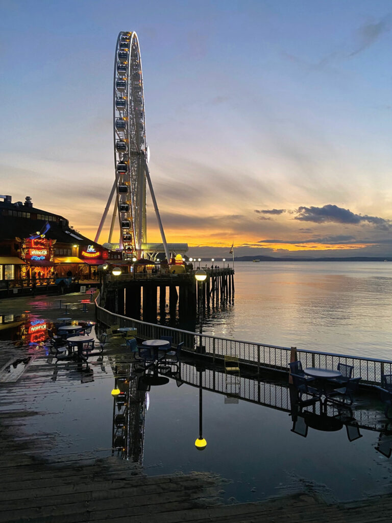 A photo of the Ferris wheel on Seattle's waterfront. The Ferris wheel sits on a pier overlooking Puget Sound. The Miner's Landing building sits to the left of it and has lit up neon signs.