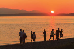 A photo of Golden Gardens Park beach at sunset. The sky is glowing orange and there is the silhouette of 8 people on the shore. The Puget Sound and Olympics Mountains are in the background.