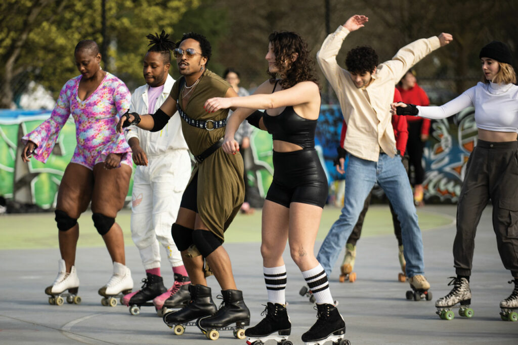 A group of people roller skate at Judkins Park. Four skaters stand in a line. One skater in the back wears a yellow shirt and waves their arms above their head.