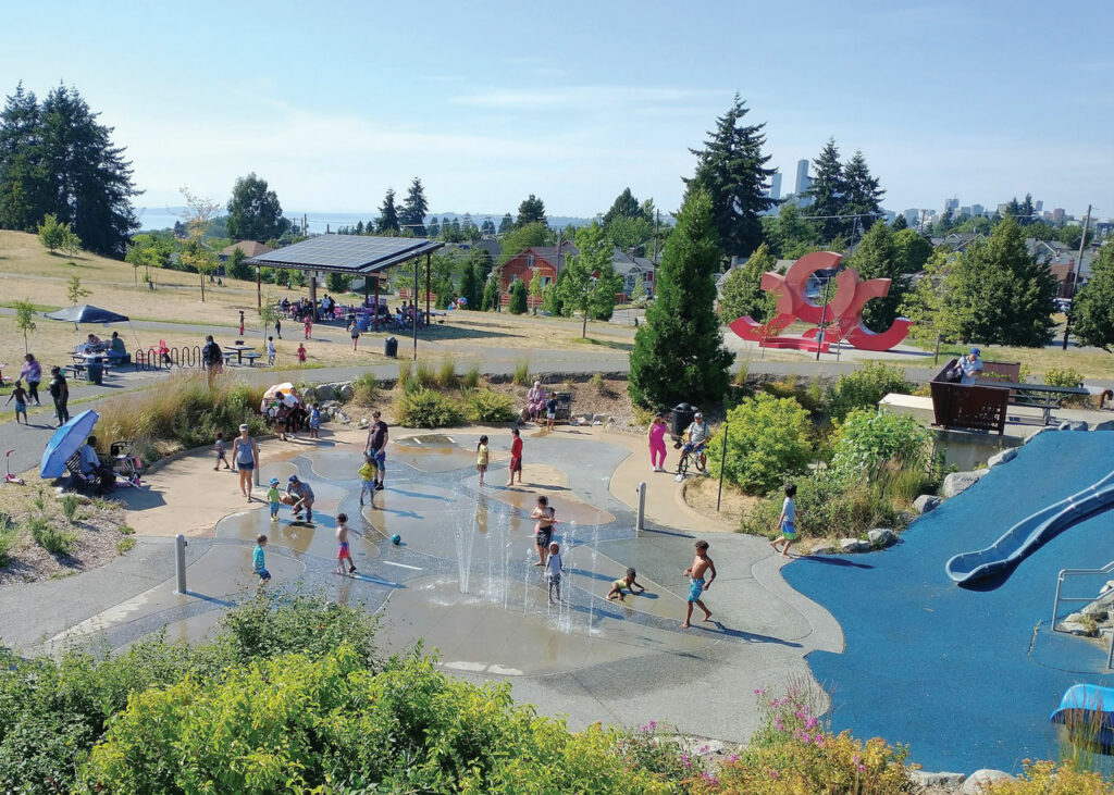 A photo of kids and families playing at a splash park. Fountains shoot water out of the ground as kids run through. A playground with a blue slide and blue ground is on the right. A field with a picnic shelter is on the left. 