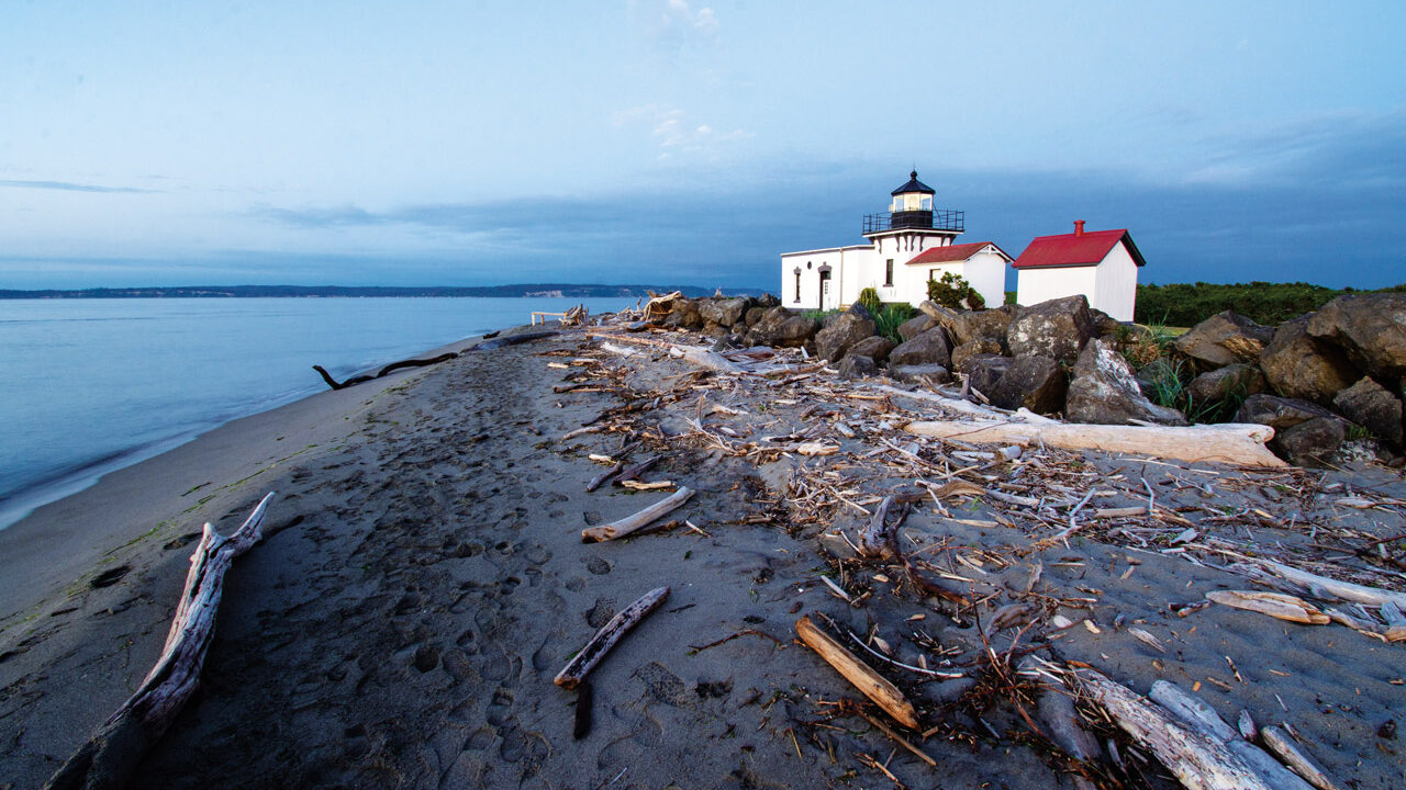 A white lighthouse with a red roof on a beach with scattered driftwood