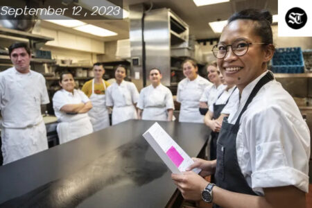 A colored women wearing a white chef outfit and black apron is smiling wearing glasses. Behind her are a group pf people wearing white around a table.