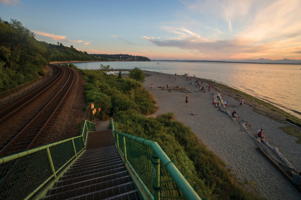 A photo of stairs heading down to the beach at Carkeek Park. The stairs have a mint green railing and lead down to a sandy beach. Puget Sound and a sunset sky are set in the background.