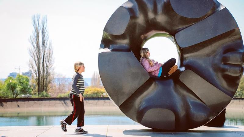 Children play among a circular black statue at the edge of a reservoir in Volunteer Park