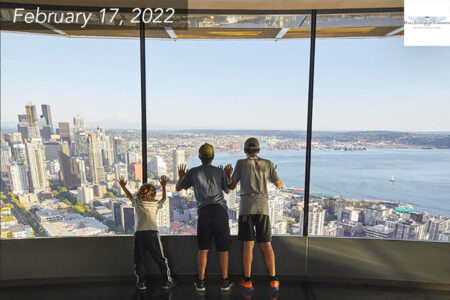 Three children look out the floor to ceiling windows of the Space Needle at the Seattle skyline.