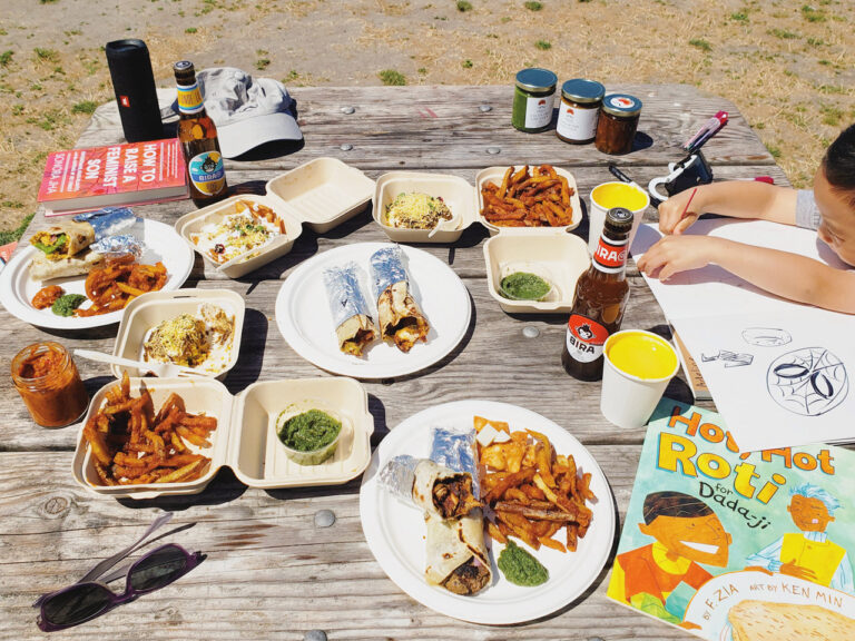 a spread of plates and takeout boxers with colorful food on a picnic table on the beach