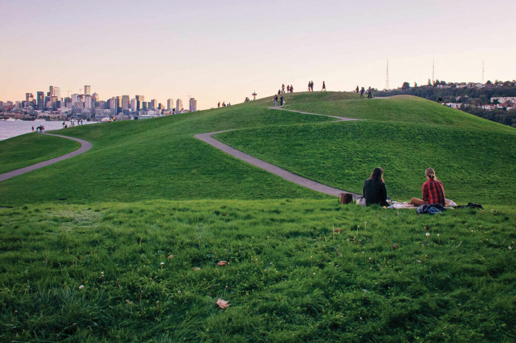 A photo of a park with a large grassy hill. The city skyline of Seattle can be seen in the background.