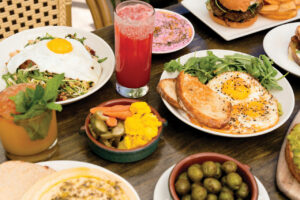 A table full of plates full of colorful food including fried eggs and toast with a side of greens, brussel sprouts, pickled vegetables, and a burger. Drinks also sit on the table. One is in a short glass and is an organge-red. The other is in a tall glass and is bright red.