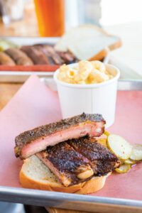 A photo of BBQ ribs set on a piece of white bread on a metal tray lined with pink paper. A white cup of mac 'n' cheese sits behind the ribs.