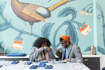 Seattle-based actor Nicholas Japaul Bernard, wearing an orange beanie, sips coffee with his friend, Siobahn in front of a decorative wallpaper design at Ben Paris.