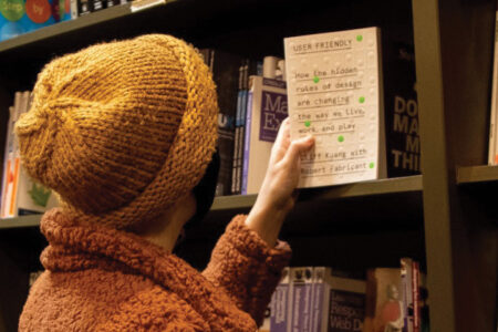 A person wearing an orange jacket and yellow beanie pulls out a book with a white cover from a bookshelf filled with books.