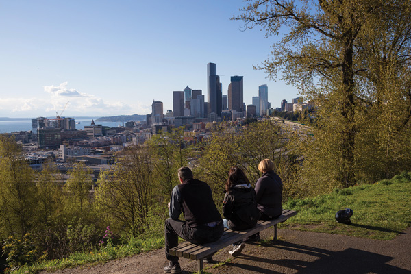 Three people sit on a bench in a green space overlooking a skyline and water in the background.