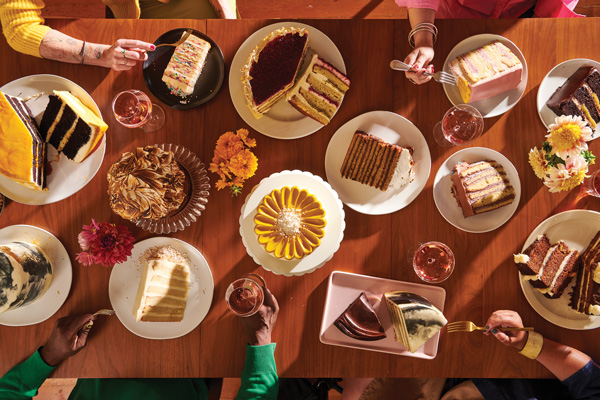 13 plates of different colored cakes sit on a wood table surrounded by flowers and drinks. Five hands reach for bites of the cake from different sides of the table.