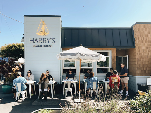 Ten people sit at white tables on a patio in front of a white, black and brick building on a sunny day.