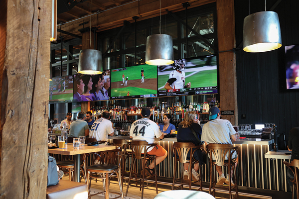 Five people sit at a bar top with three servers behind the bar. Four large screen TVs fill the background with sporting games playing.