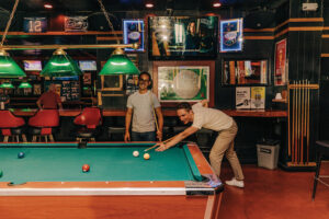 A person in a white shirt and tan pants holds a pool stick and aims to shoot a pool ball in a pub.