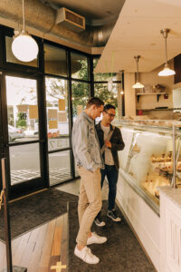 Two men wearing jean jackets stand in front of a bakery case within a shop.