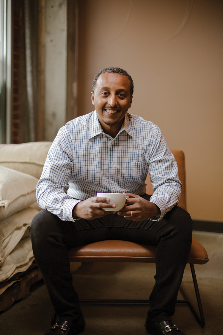A headshot of Efrem Fesaha. A person wearing a white button up shirt with blue lines and black pants. He is holding a white cup and sitting in a brown chair.