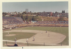 A baseball field with players in the foreground with stadium seats full of people in the background.