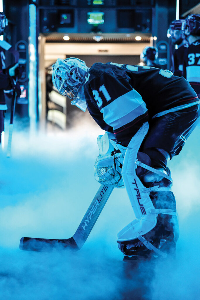A photo of Philipp Grubauer in his Kraken goalie gear holding a hockey stick. The image has a blue tone and fog swirls around the lower half of the photo.