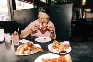 A woman in a pink shirt eats a slice of pizza off a white plate. Three additional slices of pizza and a drink surround her on a black table.